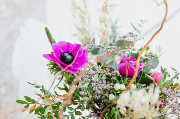Kytice s fialovými květy sasanky (anemone), staticí, bílými bobulemi třezalky, rozrazilem, listy pistácie a sušeným ovsem.