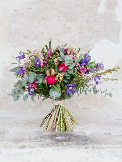 Large bouquet of seasonal flowers in pink and purple with greenery and dried bunny-tail grass and dried flax by a white wall.