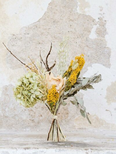 Sušená kytice uvázaná z hortenzie, mimózy, eukalyptu, lnu, ovsa a větviček kroucené vrby stojící u oprýskané bílé zdi.