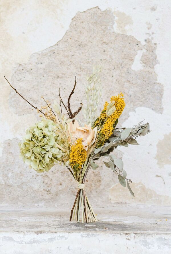Sušená kytice uvázaná z hortenzie, mimózy, eukalyptu, lnu, ovsa a větviček kroucené vrby stojící u oprýskané bílé zdi.