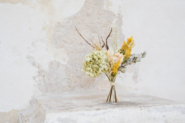 Sušená kytice stojící na bílé zdi uvázaná z eukalyptu, mimózy, hortenzie, lnu, ovsa a proutků kroucené vrby.