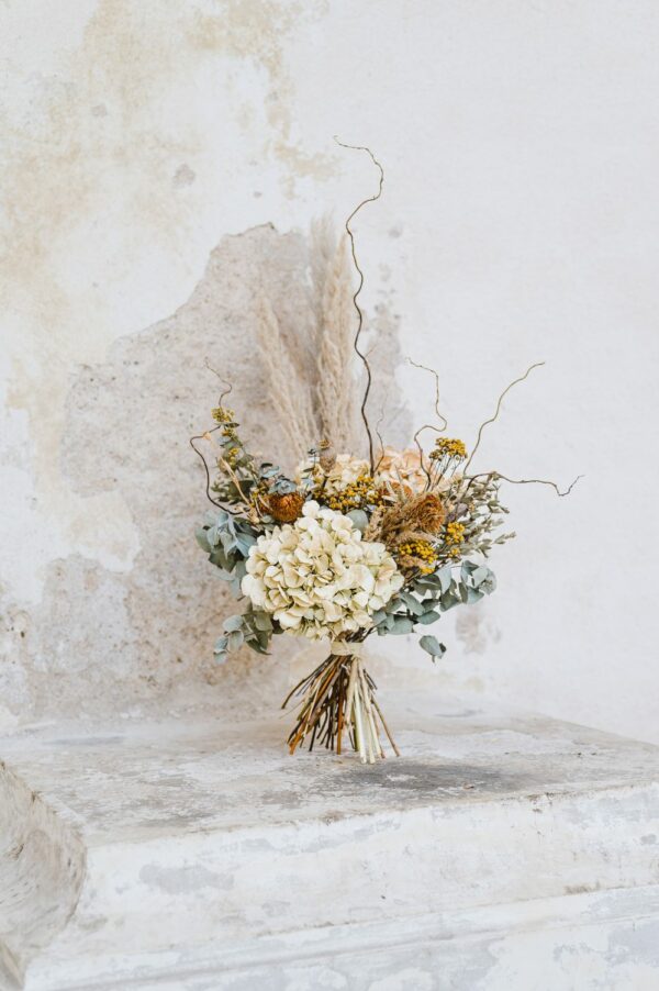 Velká kytice ze sušených květin, travin, eukalyptu a kroucené vrby stojící u bílé, oprýskané zdi.