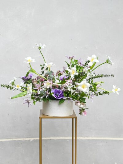 Jarní květinový box s čerstvými jarními květinami, eukalyptem a myrtou v bílém obalu na zlatém podstavci.
