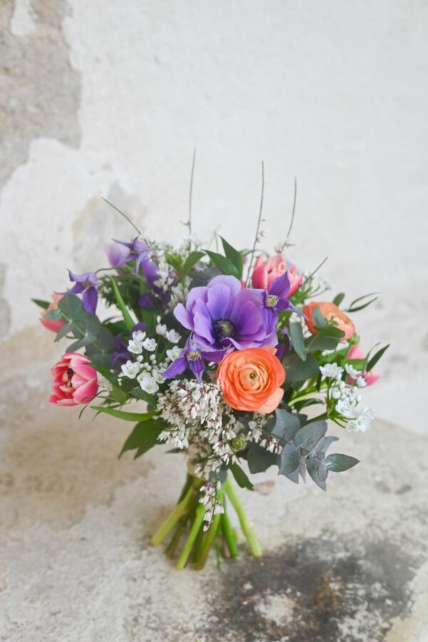 A colourful spring bouquet tied with fresh flowers in orange, purple and white and greenery stands against a chipped wall.